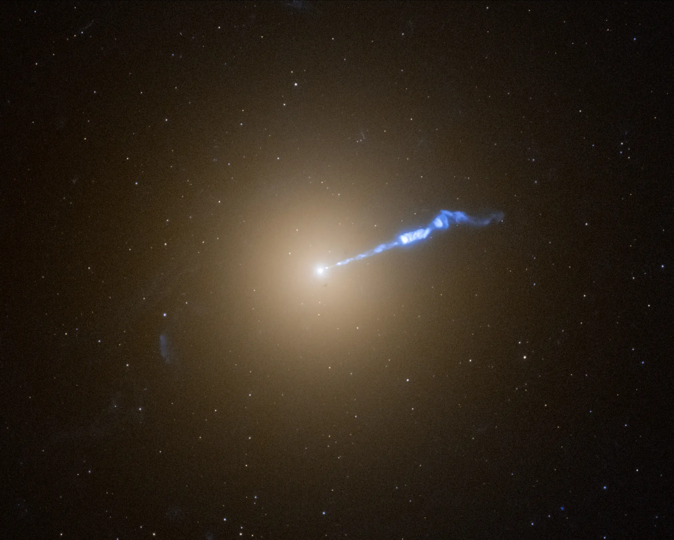 M87; sarımsı, top biçimli dev bir galaksi ve merkezinden sağa doğru uzanan ince mavi jet